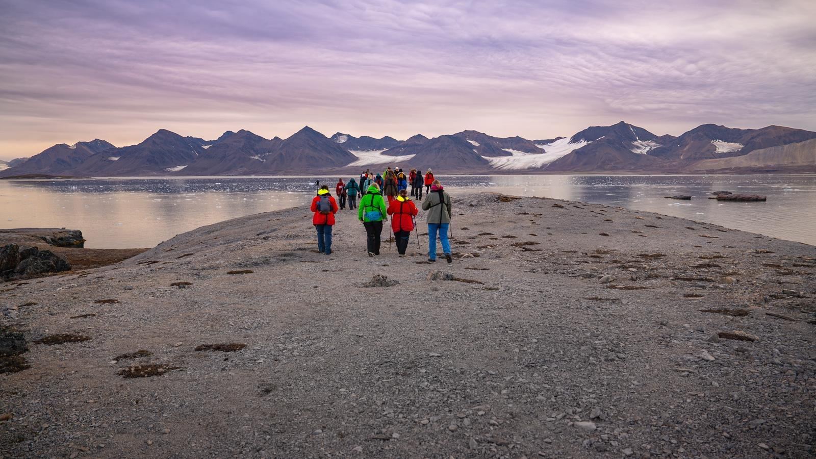 Expedition guests in bright jackets walking along a gravel spit toward Svalbard’s mountains and glacier