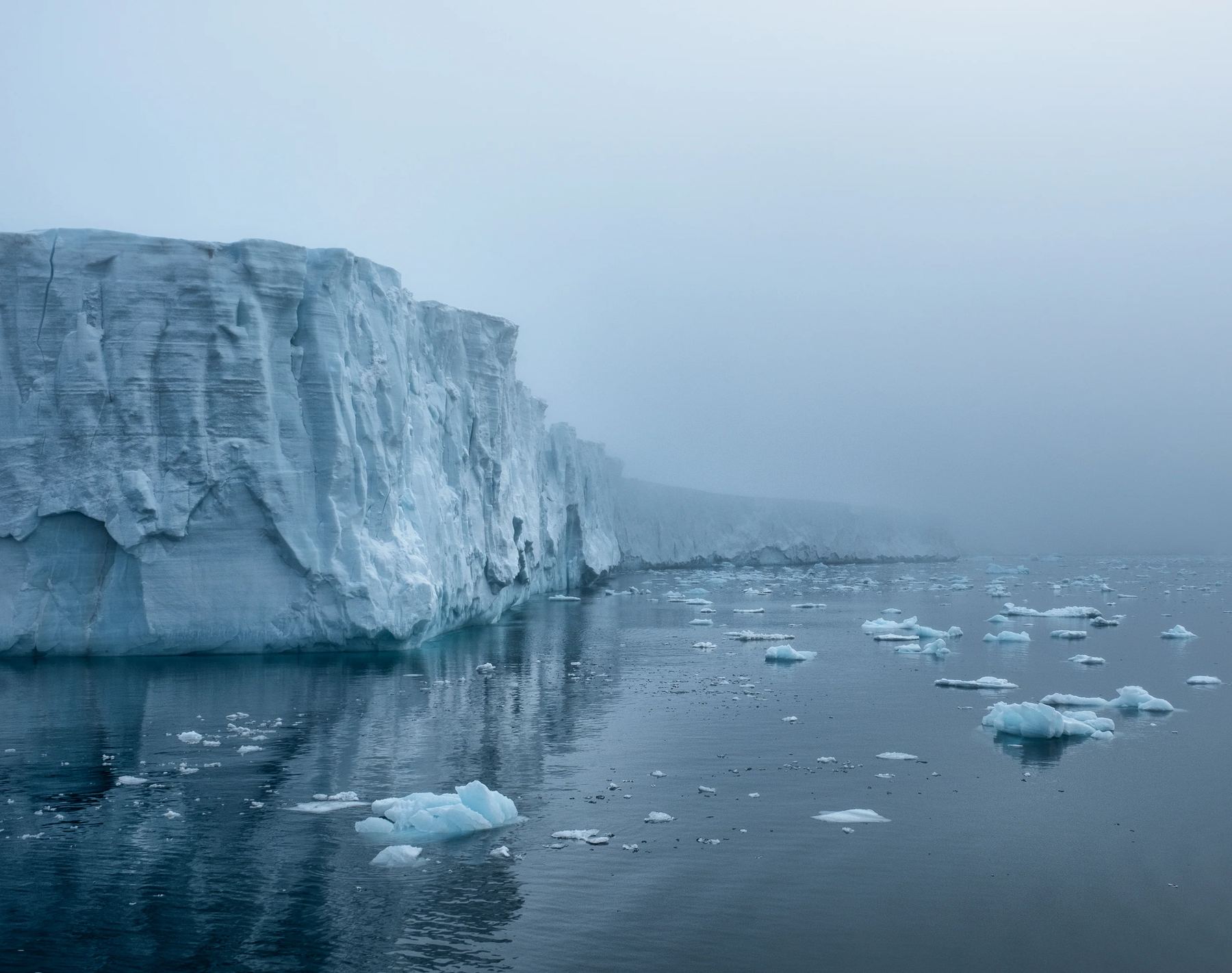 A towering blue-white glacier front rising from calm Arctic water, with floating ice and fog