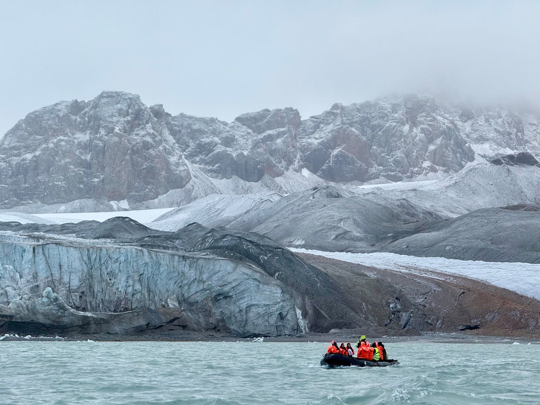 A small zodiac carrying guests in orange suits in front of a glacier and snow-dusted mountains in Svalbard